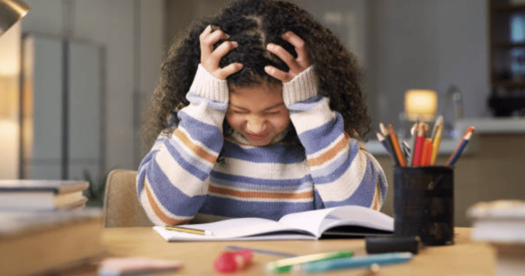 child with her hands in her hair sitting at a table with a book in front of her - toxic stress